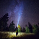 Person standing in a field at night, using a flashlight to illuminate the surroundings while gazing at a starry sky, highlighting the beauty of nature. Ideal for showcasing Michigan web design themes focused on outdoor adventures and exploration.