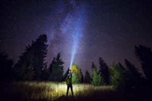 Person standing in a field at night, using a flashlight to illuminate the surroundings while gazing at a starry sky, highlighting the beauty of nature. Ideal for showcasing Michigan web design themes focused on outdoor adventures and exploration.